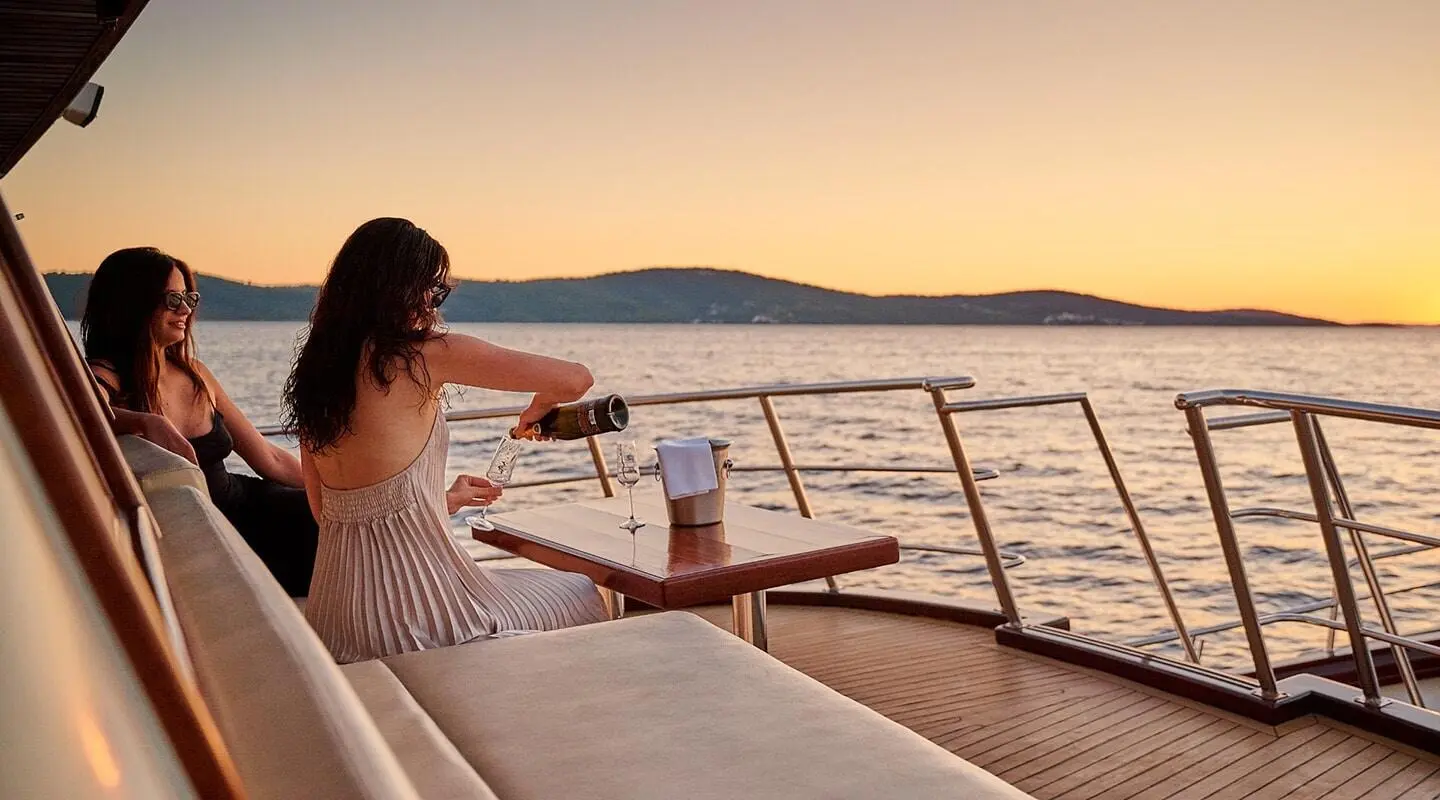 Luna yacht sunset lounge: woman pours champagne on a teak deck with sea horizon