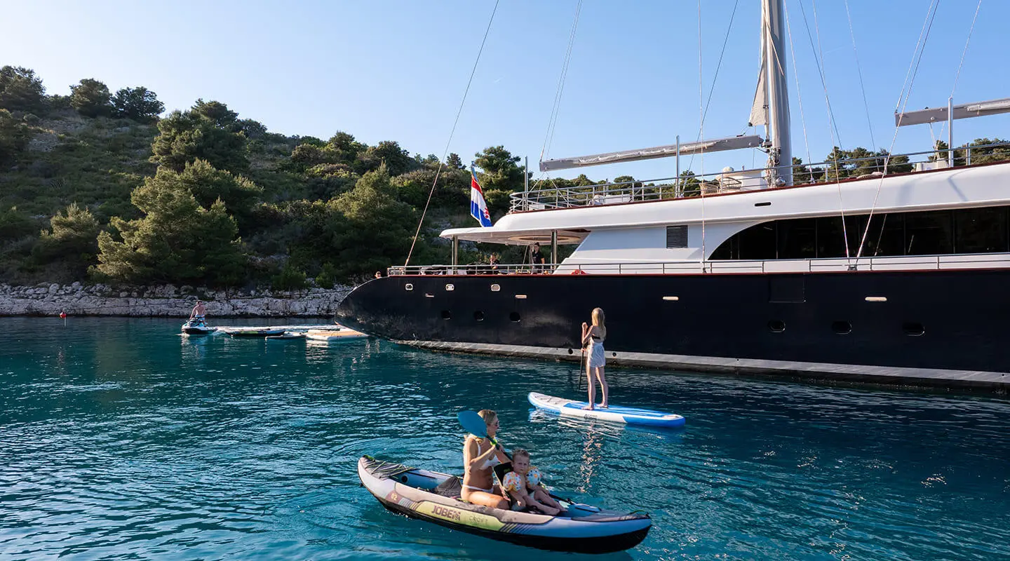 Two people on a small boat near a dark yacht named Nocturno anchored by rocky shore