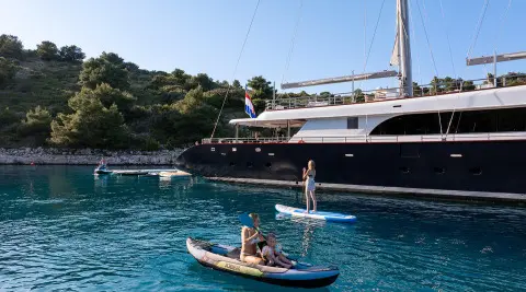 Two people on a small boat near a dark yacht named Nocturno anchored by rocky shore