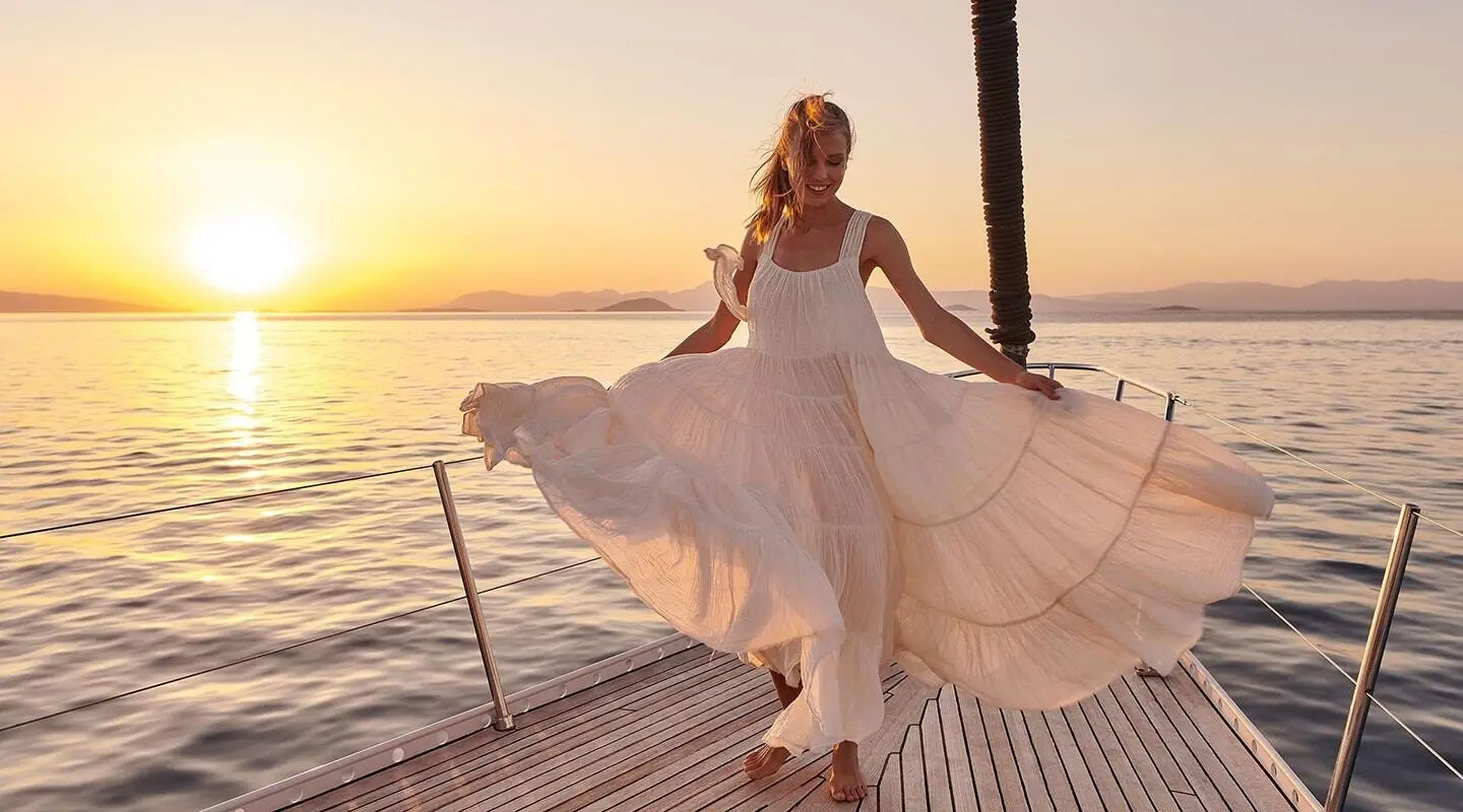 Woman in flowing dress on yacht deck at sunset with sail and railing