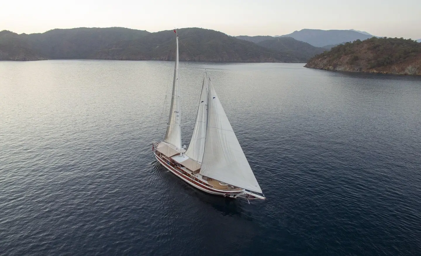 Kayhan Kaptan yacht sailing on calm sea near rocky coastline with sails raised
