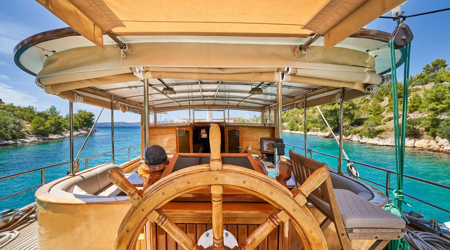 Libra yacht wheel on deck with sunlit seating and white sails behind