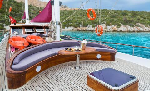 Luxurious wooden deck with curved seating and table, orange life rafts, open sea and rocky shore in background