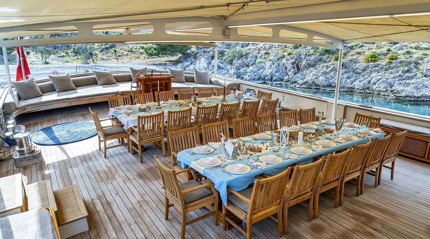 Tersane 8 yacht dining area on deck with long table set for guests against rocky shore