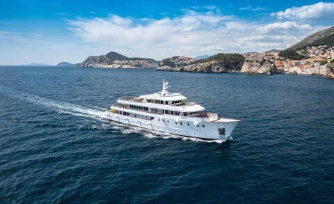 White Yolo motor yacht cruising on open sea with rocky coastline and town in the background