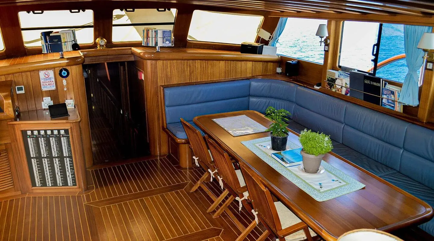 Inside view of Enderim A yacht: teak dining area with table, bench seating, and potted plant, afternoon light