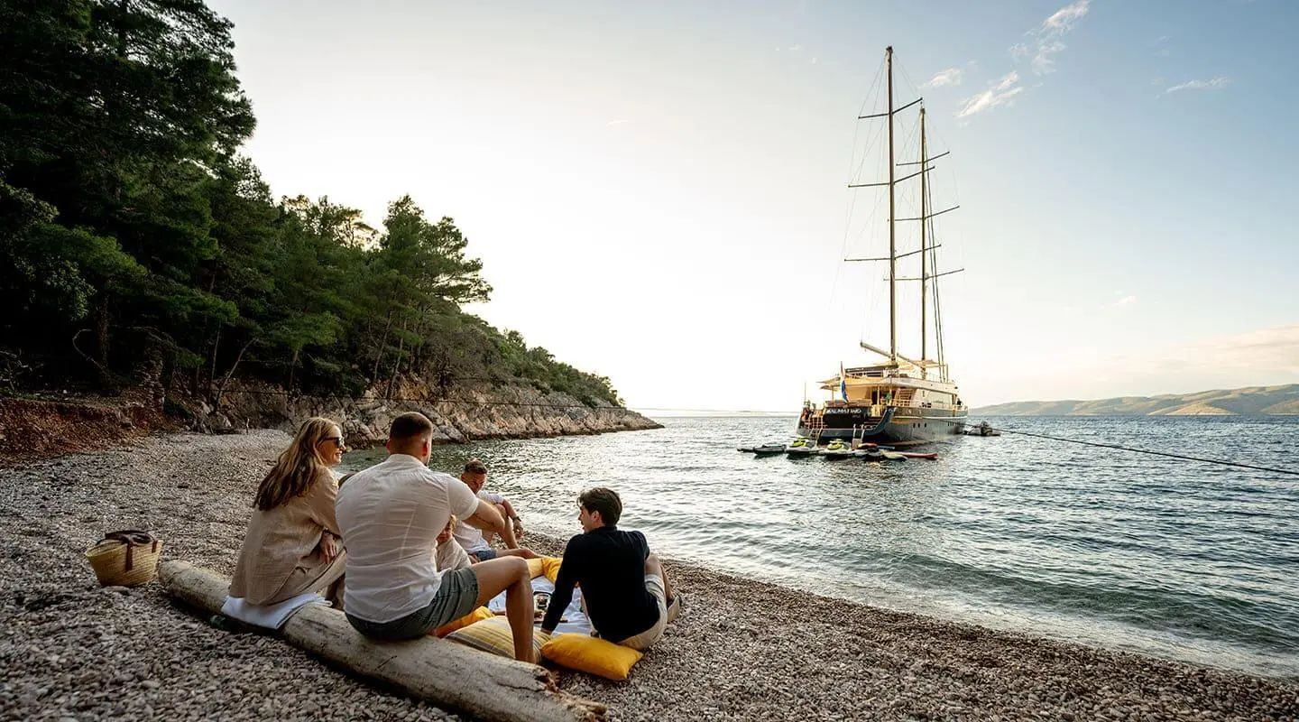 Dalmatino yacht anchored near rocky shore at sunset with people on pebbled beach