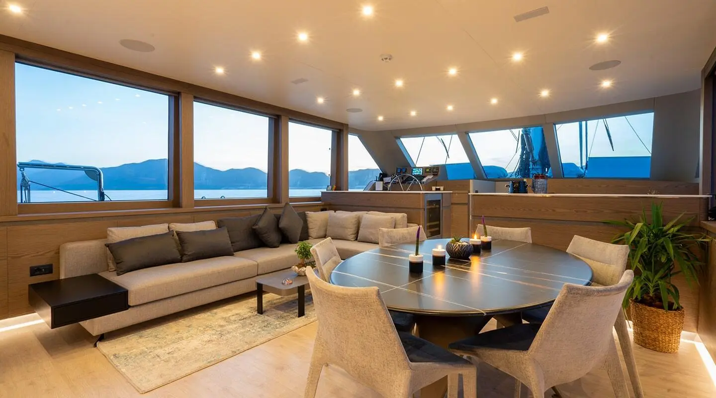 Interior of a yacht saloon with beige seating, wooden table, large windows, calm sea view