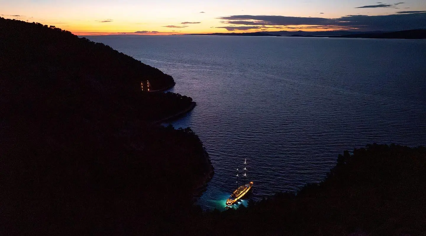 Dalmatino yacht anchored in a secluded cove at dusk with illuminated deck lights