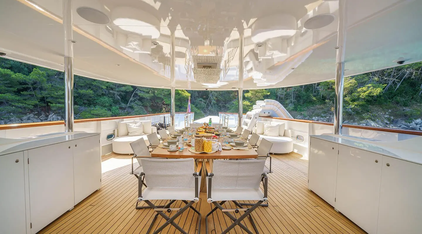Maia yacht open deck with a long dining table, white seating and cabinets