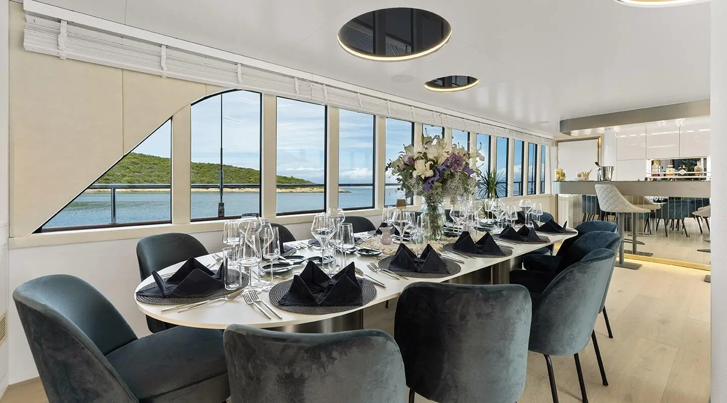 Interior dining area aboard Maxita yacht with long table set for guests, large windows, and elegant seating