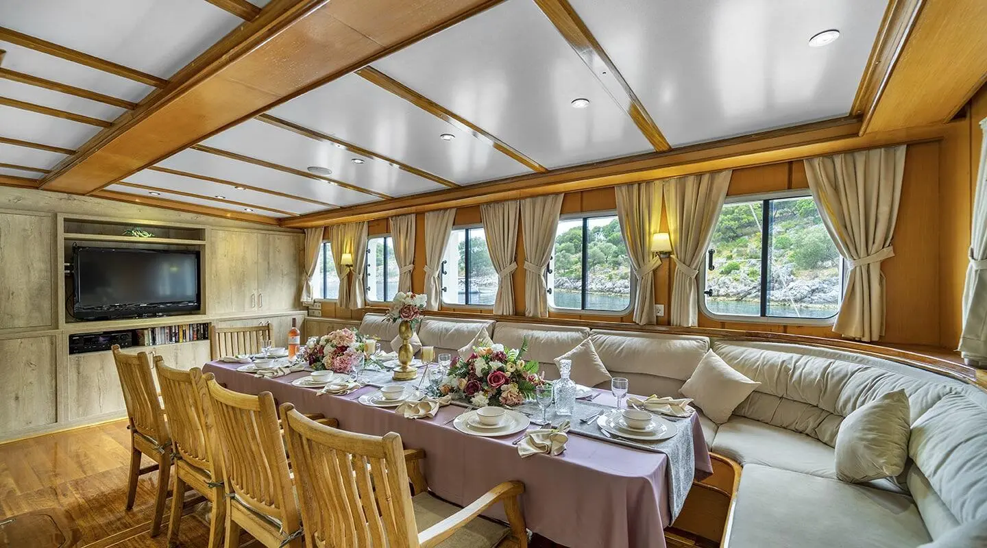 Long dining table with wooden chairs on a yacht interior, with windows and white seating
