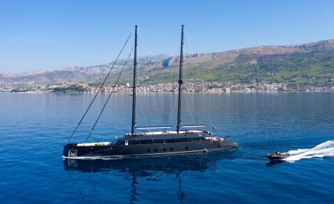 Black-hulled sailing yacht cruising on calm blue water with a coastal town and mountains in the background