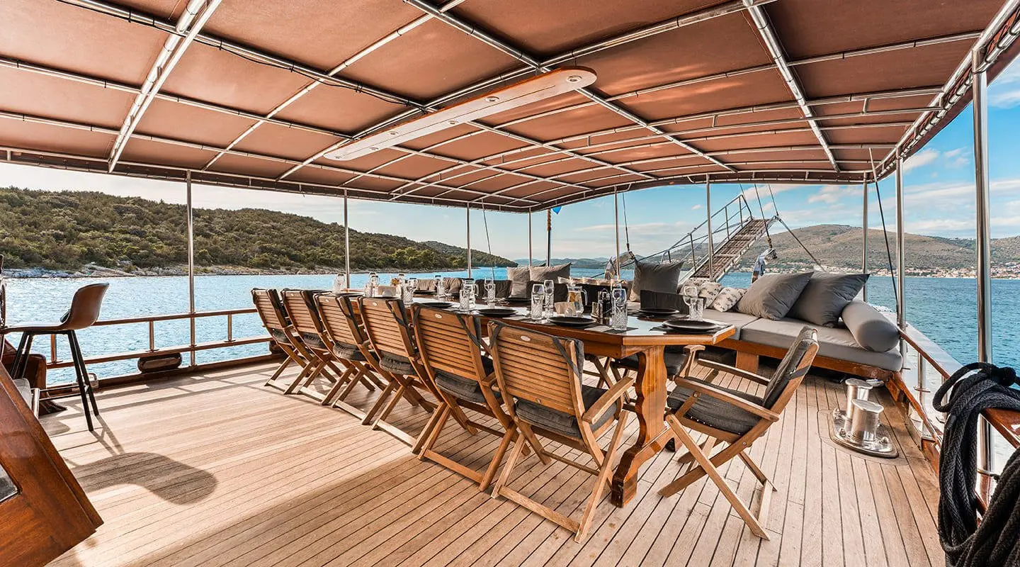 Sunlit yacht deck dining area with wooden table and chairs under a canopy