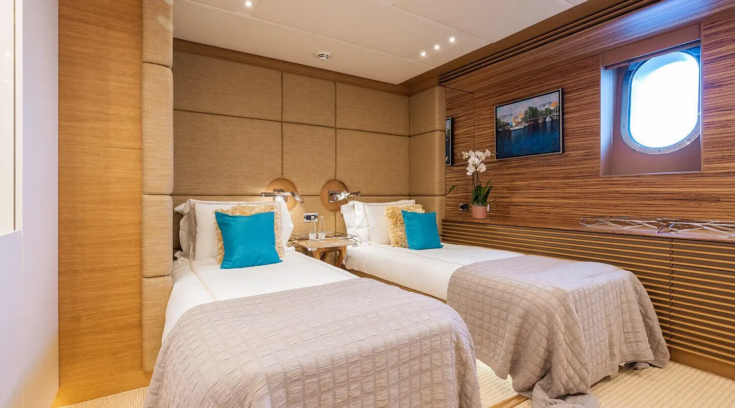 Two beige upholstered beds with teal and white pillows in a yacht cabin