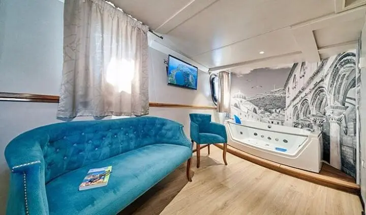 Casablanca yacht cabin with teal sofa, desk, TV, and large window draped curtains