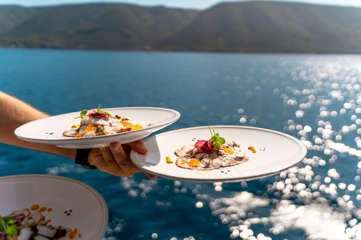 Two plates of food held over sparkling blue water with a distant coastline