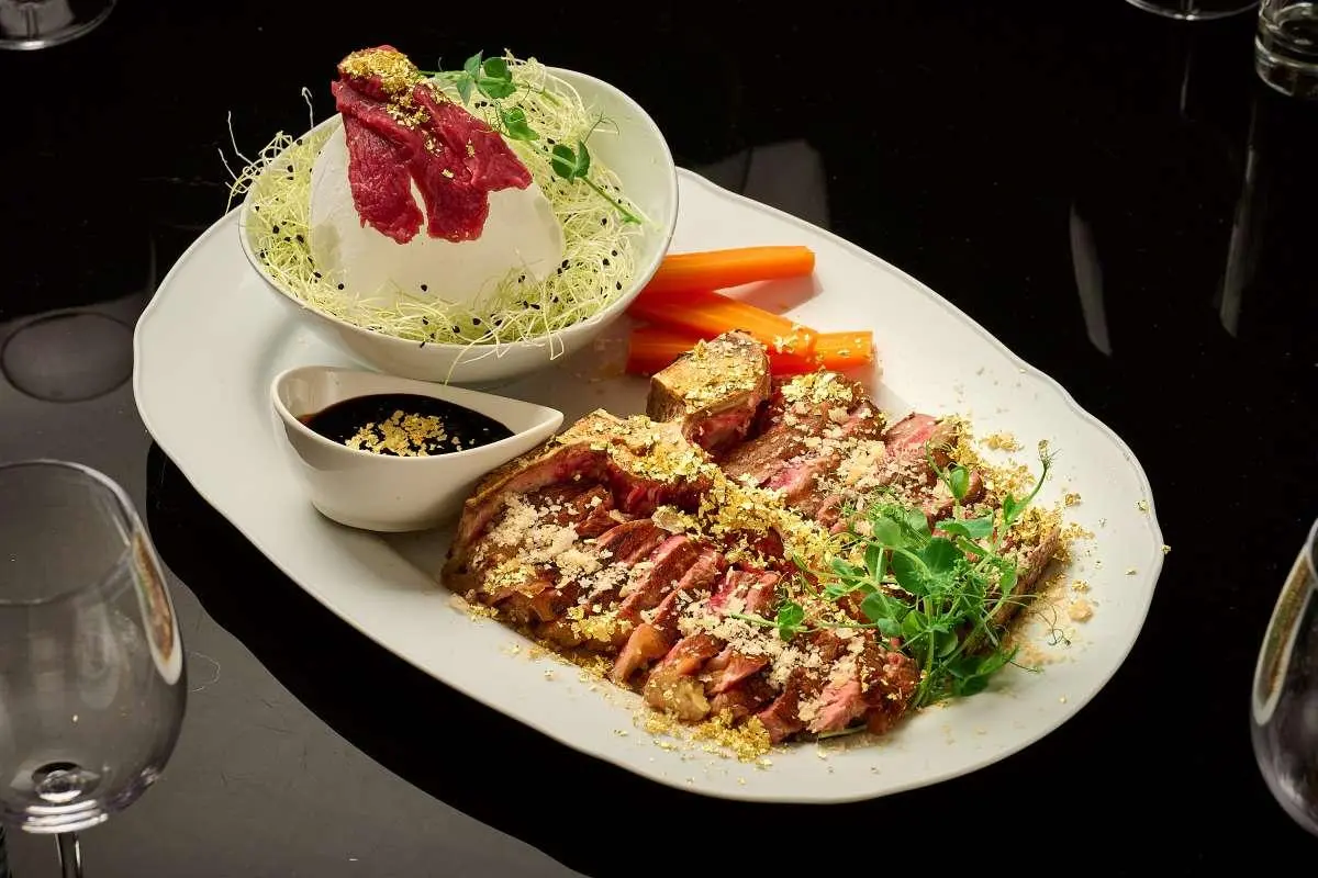 Plate of grilled meat slices with sauce and a small side bowl on a dark table