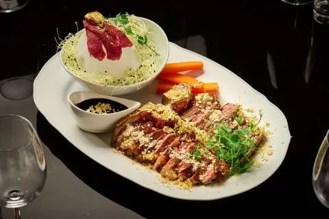 Plate of grilled meat slices with sauce and a small side bowl on a dark table