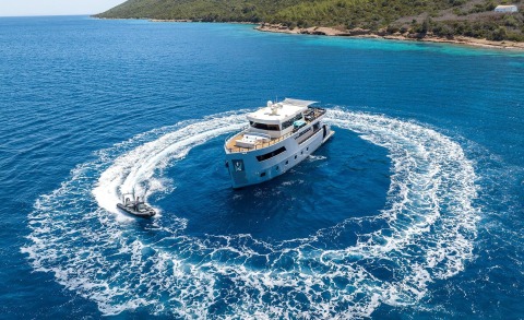 White motor yacht Calm Down makes a circular turn in blue sea with coastline in the background and wake trails