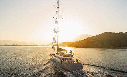 Sailing yacht on open sea at sunset with tall mast and wake behind.
