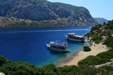 Two passenger boats anchored in clear blue water beside a rocky, green shoreline
