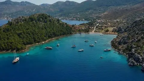 Turquoise bay with anchored sailboats near a forested coastline and hills