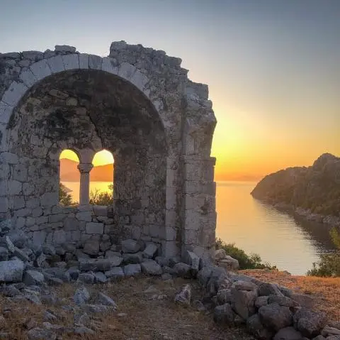 Stone arched ruin window framing a sunset over calm sea and rocky coastline.