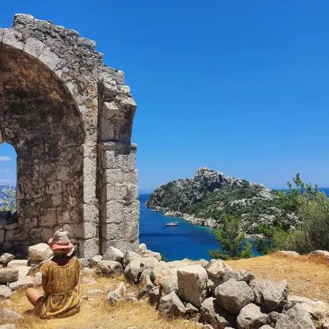 Woman in a hat sitting by an ancient stone arch, blue sea and rocky island view