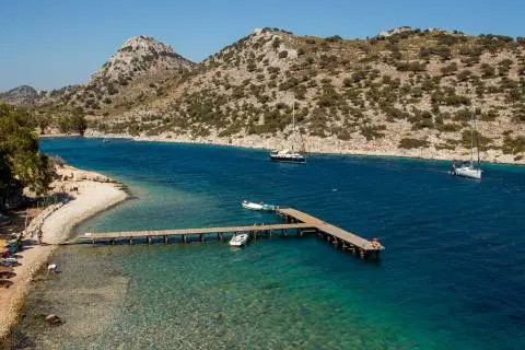 Wooden pier extending into clear turquoise water, anchored sailboats and rocky hills