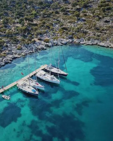 Four sailing yachts moored at a wooden pier beside rocky shore and turquoise clear water