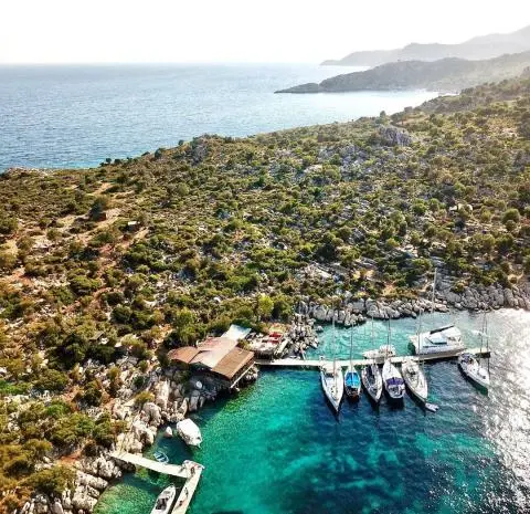 Sailboats moored at a small dock in a turquoise cove beside rocky, shrub-covered coastline