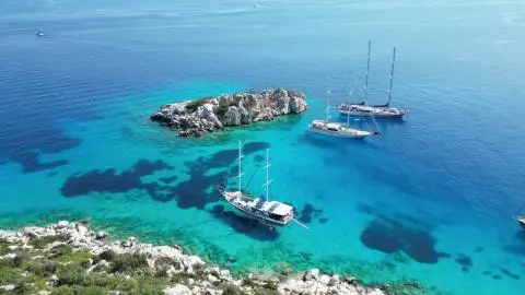 Two-masted gulet and two yachts anchored by a rocky islet in turquoise sea