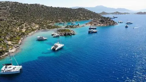 Yachts anchored in a shallow turquoise cove with rocky, pine‑covered shoreline