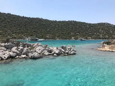 Turquoise bay with rocky shore, people wading and two anchored boats near wooded hillside