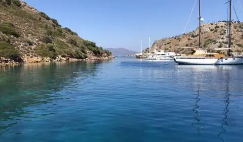Calm cove with clear blue water flanked by rocky shores and a distant sailboat