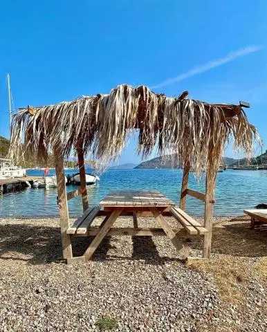 Thatch-covered wooden picnic table on a pebble beach by a clear blue bay with docked boats