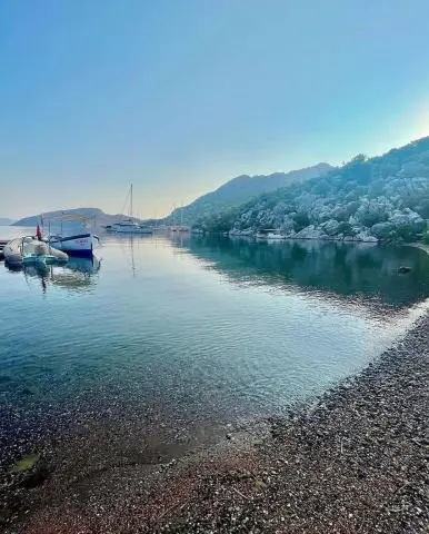 Calm cove with pebbled shore, small boats, and distant sailboats on blue water