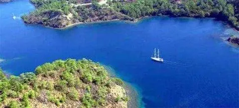 White two-masted sailing yacht in a deep-blue bay surrounded by pine-covered coves