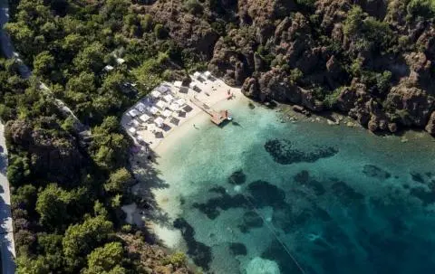 A turquoise bay with a sandy beach and a small pier along rocky cliffs