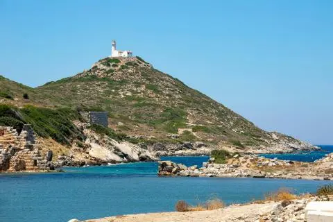 White lighthouse on a hill with scrubby vegetation and clear blue sea in foreground