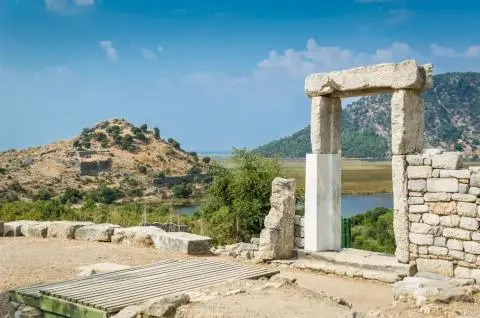Stone-columned ancient gate ruins with a lake and shrub-covered hills visible behind