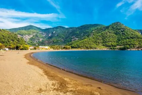 Sandy shoreline curving along calm blue water with green forested hills behind