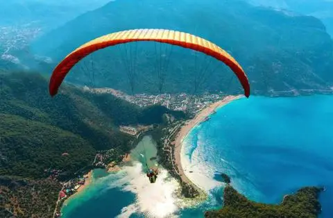 Person paragliding under an orange wing over a bay and turquoise sea in an aerial view