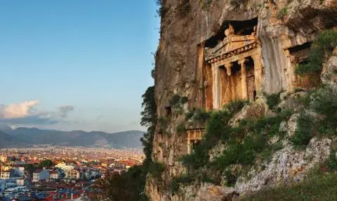 Ancient rock-cut tomb facade on cliff with cityscape and distant mountains under clear sky
