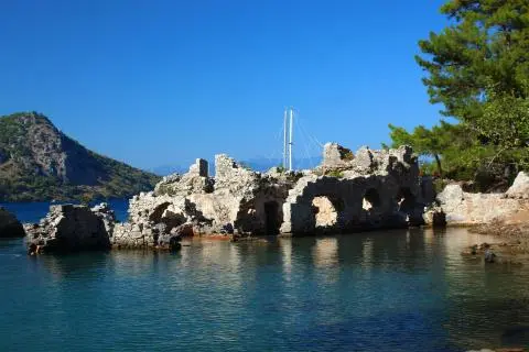 Stone-arched ruins beside calm blue sea with a sailboat mast nearby