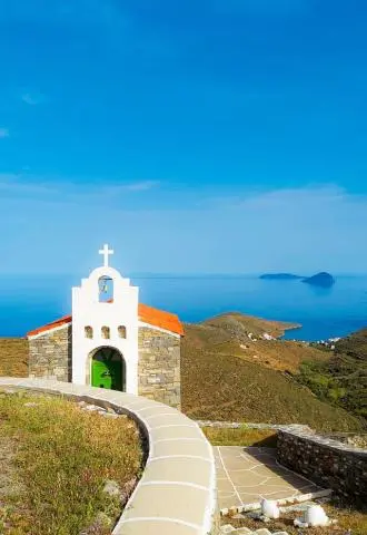 Small white chapel with green door and bell overlooking hillside and calm blue sea