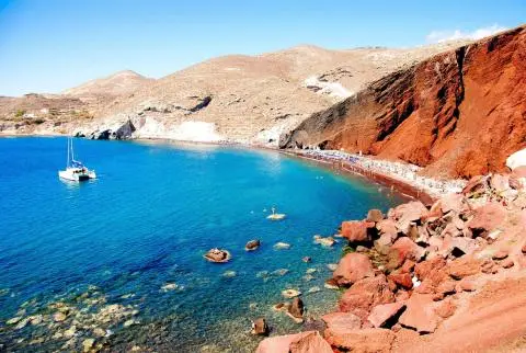 White sailing yacht anchored in turquoise bay beside red volcanic cliffs and crowded beach