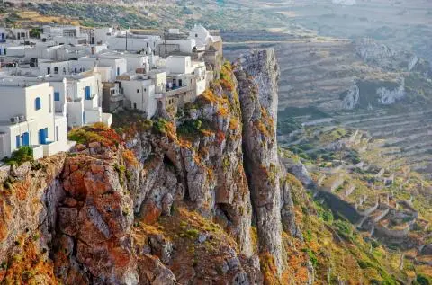 White houses with blue shutters perched on a cliff beside terraced rocky hills and winding stone walls