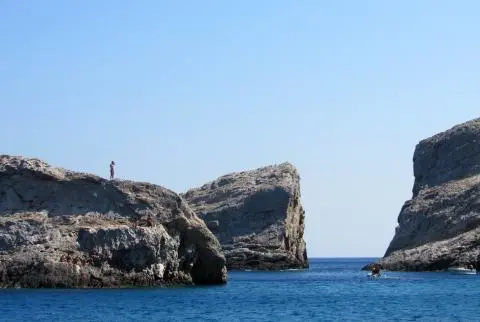Rocky cliffs with a person standing on the left promontory and small boats in deep blue sea